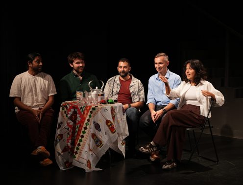 Five improvisers seated around a table with a karak kettle before a show